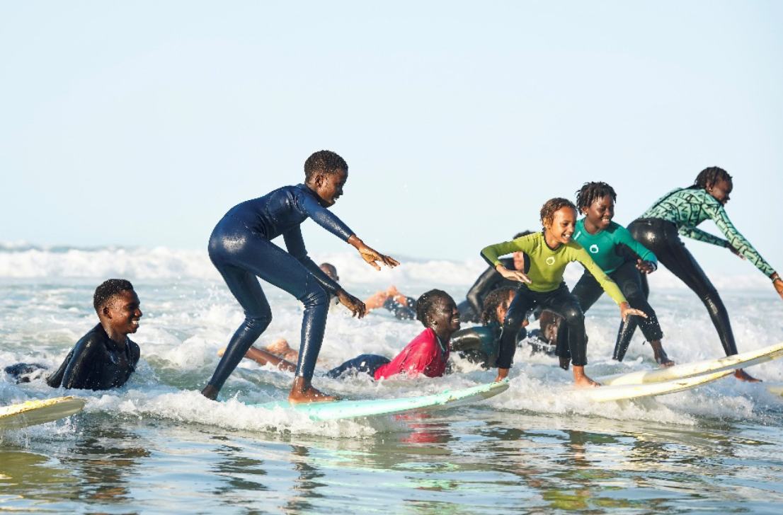 Jugendliche surfen in Neoprenanzügen auf kleinen Wellen.